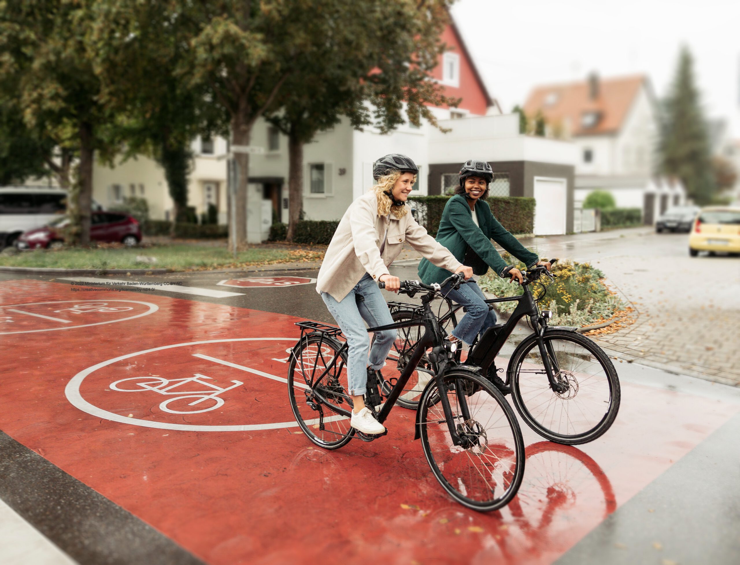 Umgebauter_Strassenraum_Radfahrerinnen_edited Zwei Radfahrerinnen fahren über eine breite Fahradmarkierung im Kreuzungsbereich einer Straße.